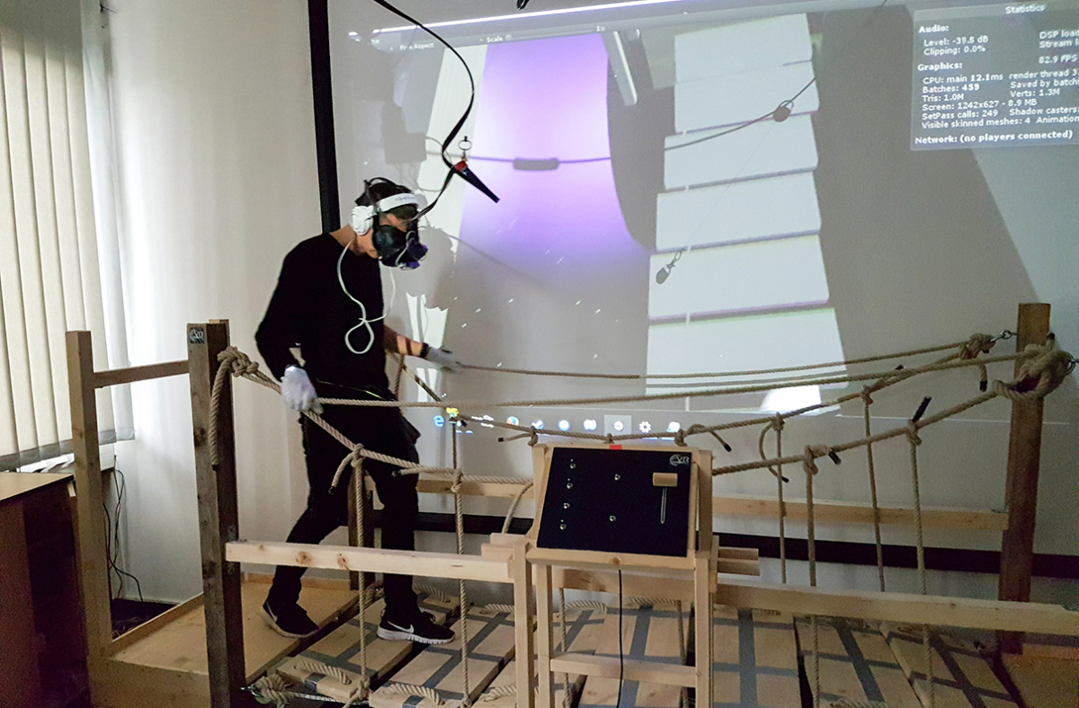 Ein Mensch, der eine VR-Brille trägt und in einem Forschungslabor über eine Hängebrücke läuft. Im Hintergrund sieht man das Bild aus der VR-Brille über einen Beamer dargestellt. Auf dem Bild sind graue Blöcke in der Luft zu sehen, über die die Person in VR läuft.