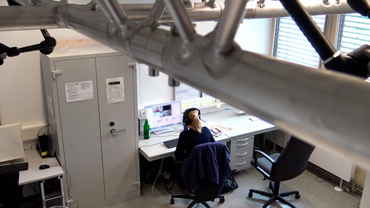 Die Kamera filmt an einer Deckenaufhängung vorbei auf einen Arbeitsplatz. Dort sitzt eine Person an einem Computer.