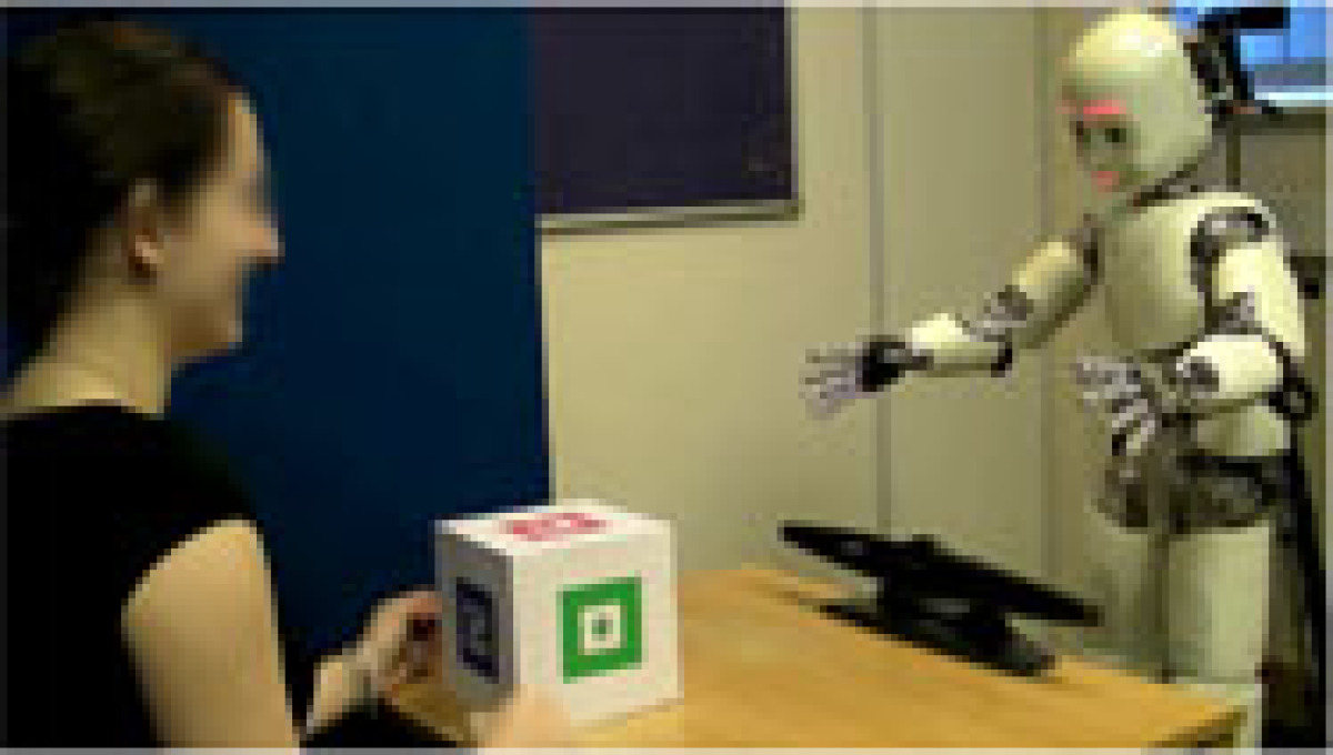 A Person sits on a table across from a robot, holding a white cube in their hands.