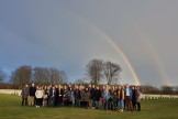 The picture shows a group of students and teachers standing outside in a green field beneath a rainbow.