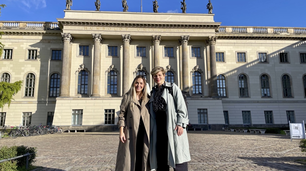 Two smiling white women stand in front of a large building with columns and the name Humboldt Universitaet on it, with a blue sky in the background