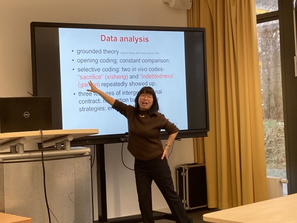 Photo of an Asian woman speaking and pointing to a powerpoint presentation with the text 