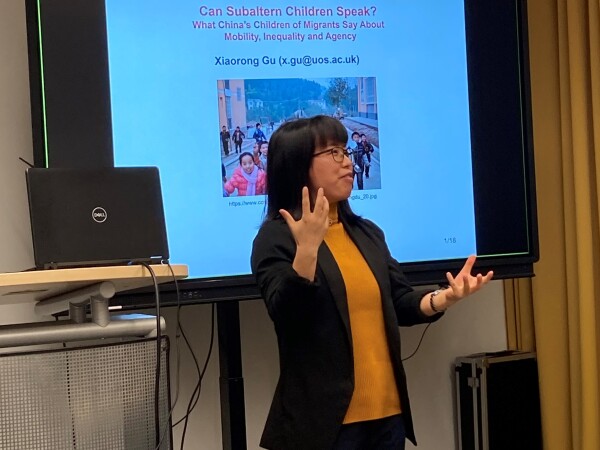 Photo of an Asian woman speaking and gesturing with her hands in front of a powerpoint presentation with the text 