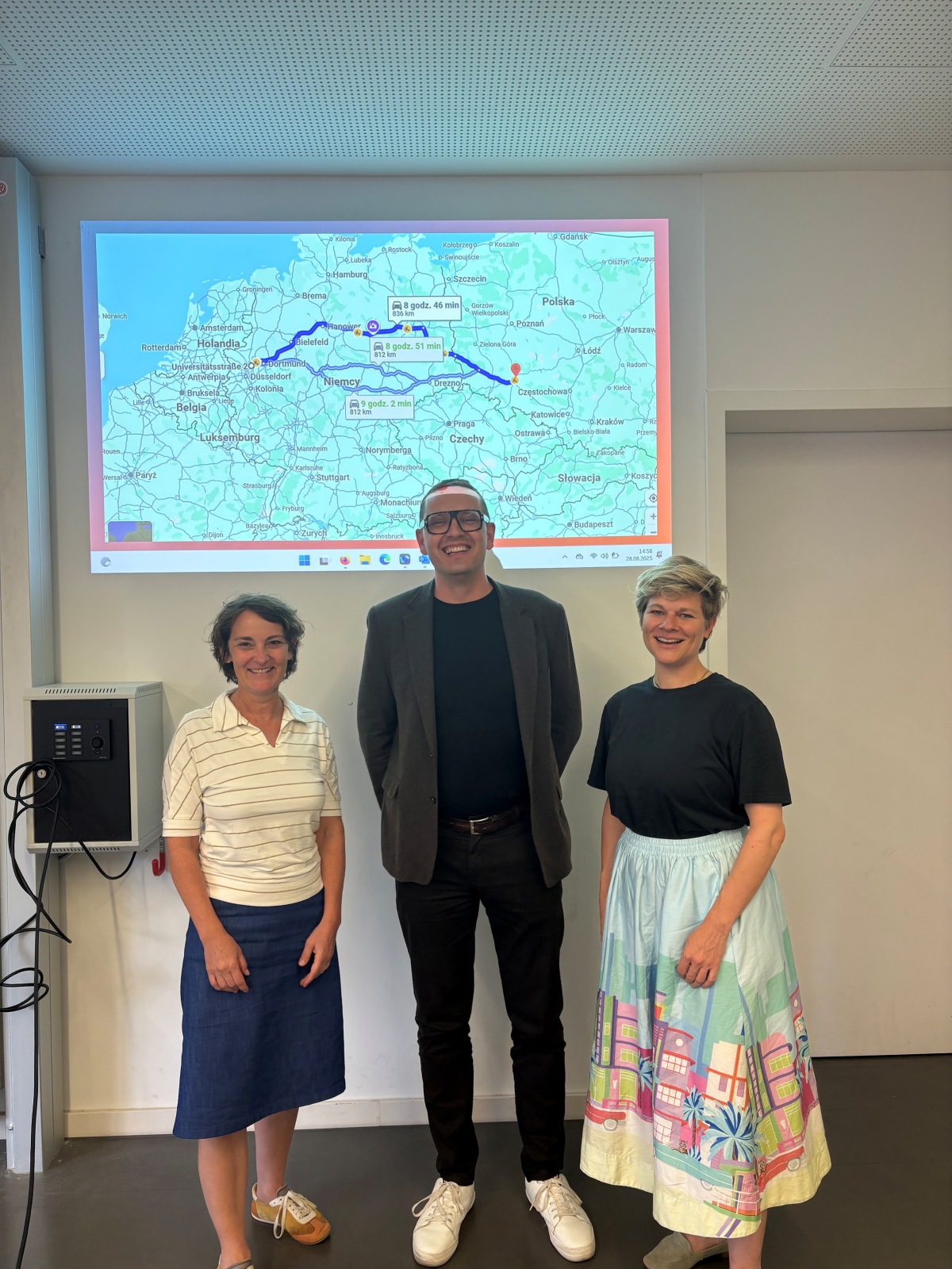 Photo of a white woman, a taller white man, and second white woman, smiling at the camera and standing in front of a projection showing the driving distance between a city in Germany and a city in Poland