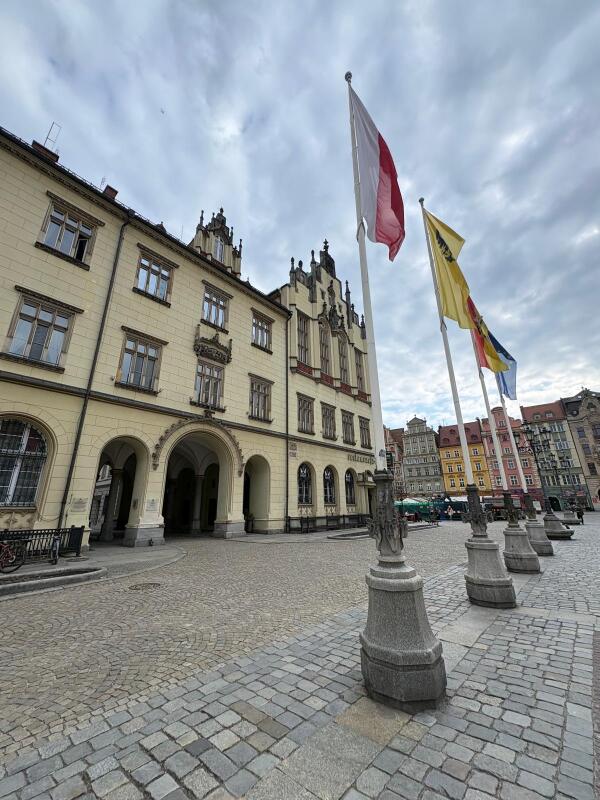 Picture of the front of an ornate yellow building. In front of the building are four flags mounted on stone bases