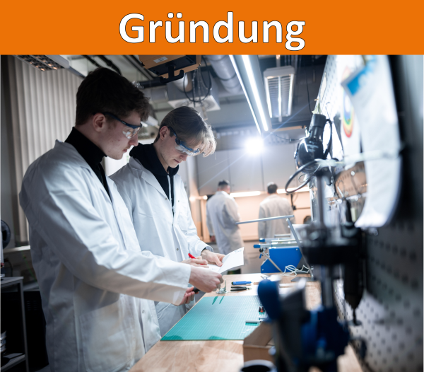 Ein oranger Balken mit der Überschrift Gründung. Darunter ein Bild von zwei Personen die mit Laborkittel und Schutzbrille an einer Werkbank arbeiten.