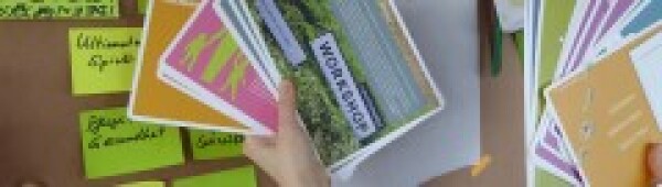 Hand hält eine Karte, auf der Workshop steht, vor eine Pinnwand mit Post-its