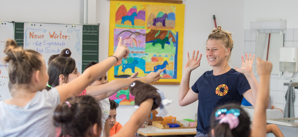Mehrere Kinder stehen im Klassenraum um die Seminarleitung und melden sich. Die Seminarleitung hebt lachend die Hände.