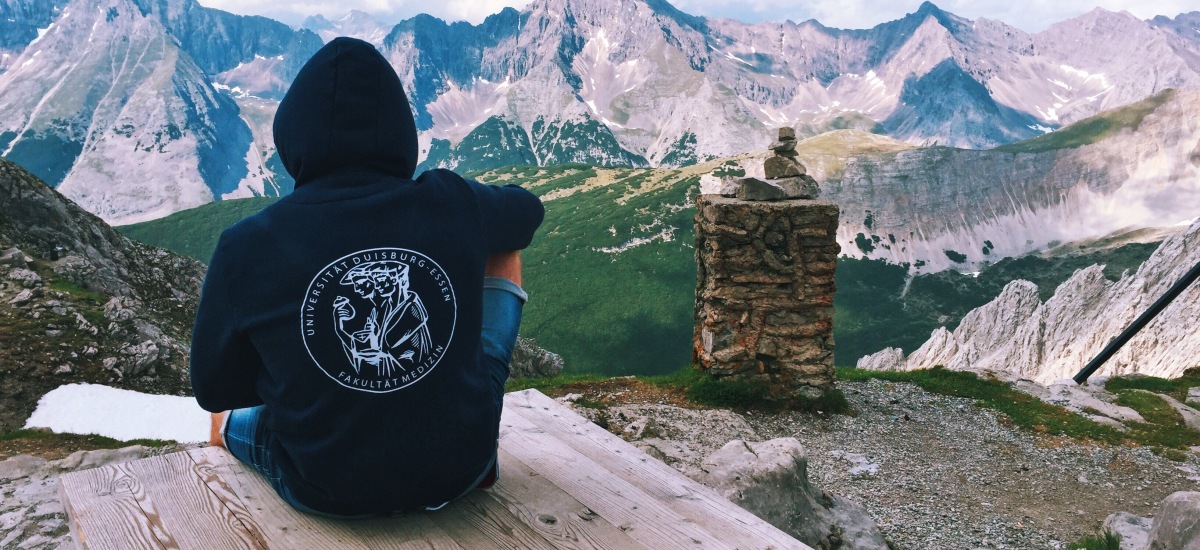 UDE Student sitzt mit UDE Hoodie auf einem Berg und genießt die Aussicht der Berge.