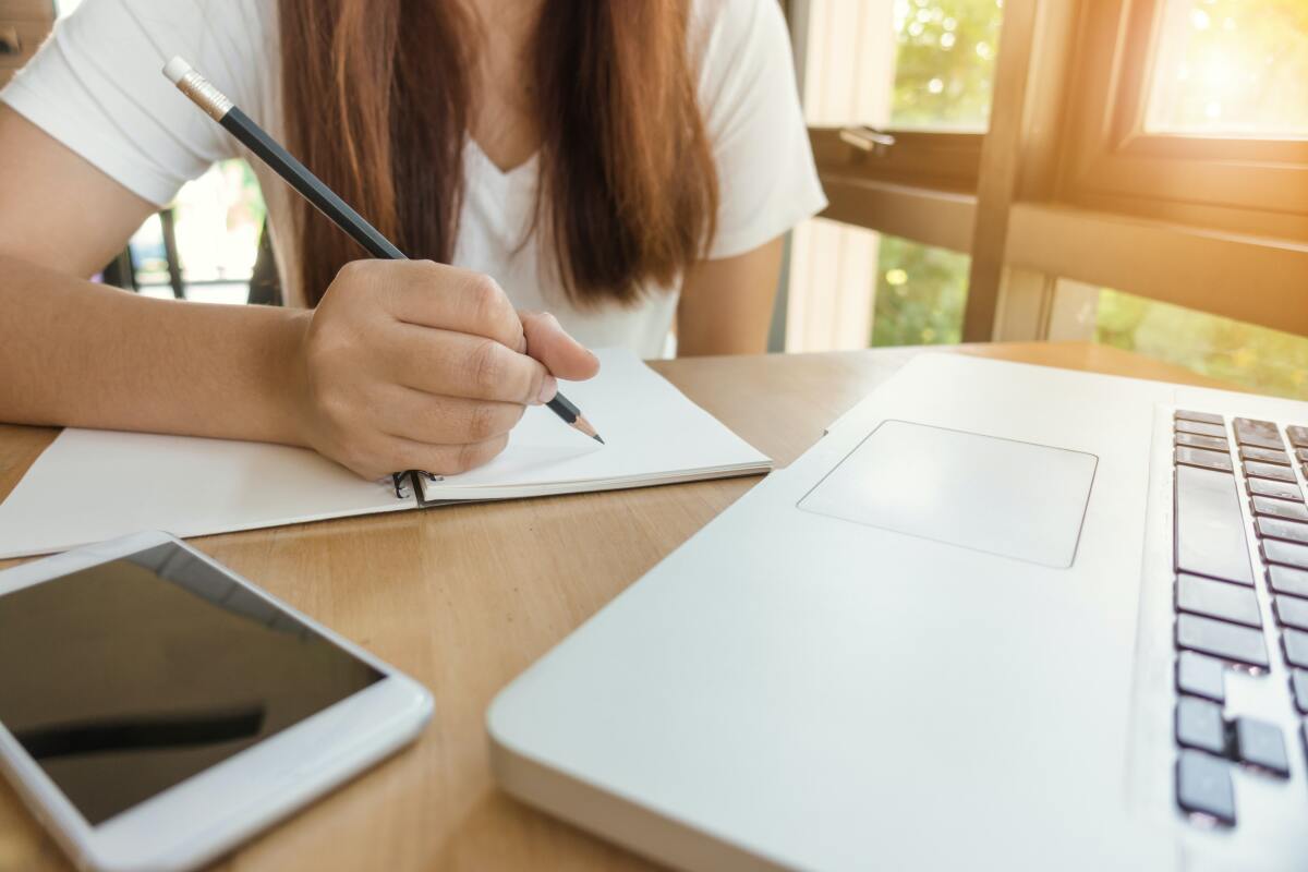 Eine Frau, die vor einem Laptop sitzt und etwas in ein Notizbuch notiert.