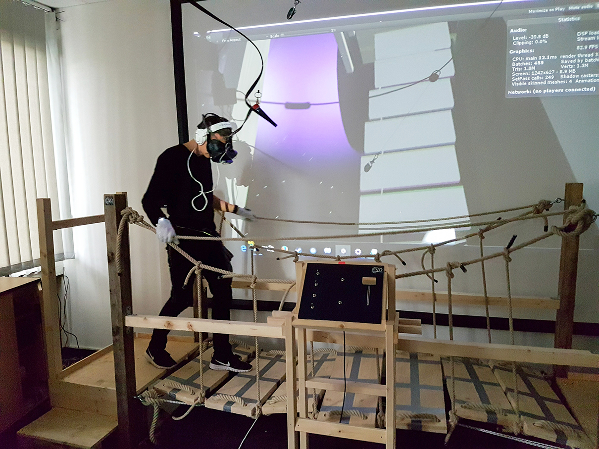 Ein Mensch, der eine VR-Brille trägt und in einem Forschungslabor über eine Hängebrücke läuft. Im Hintergrund sieht man das Bild aus der VR-Brille über einen Beamer dargestellt. Auf dem Bild sind graue Blöcke in der Luft zu sehen, über die die Person in VR läuft.