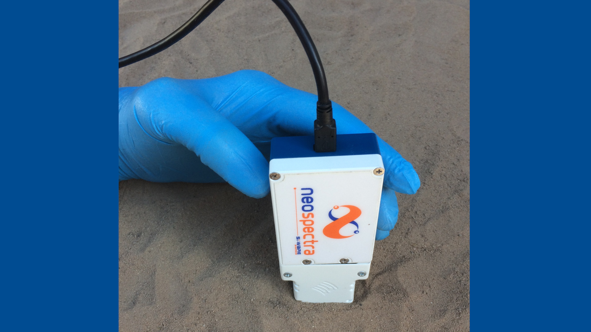 Eine Hand im blauen Handschuh hält ein Messgerät etwa in DIN A6-Format in Sandboden.