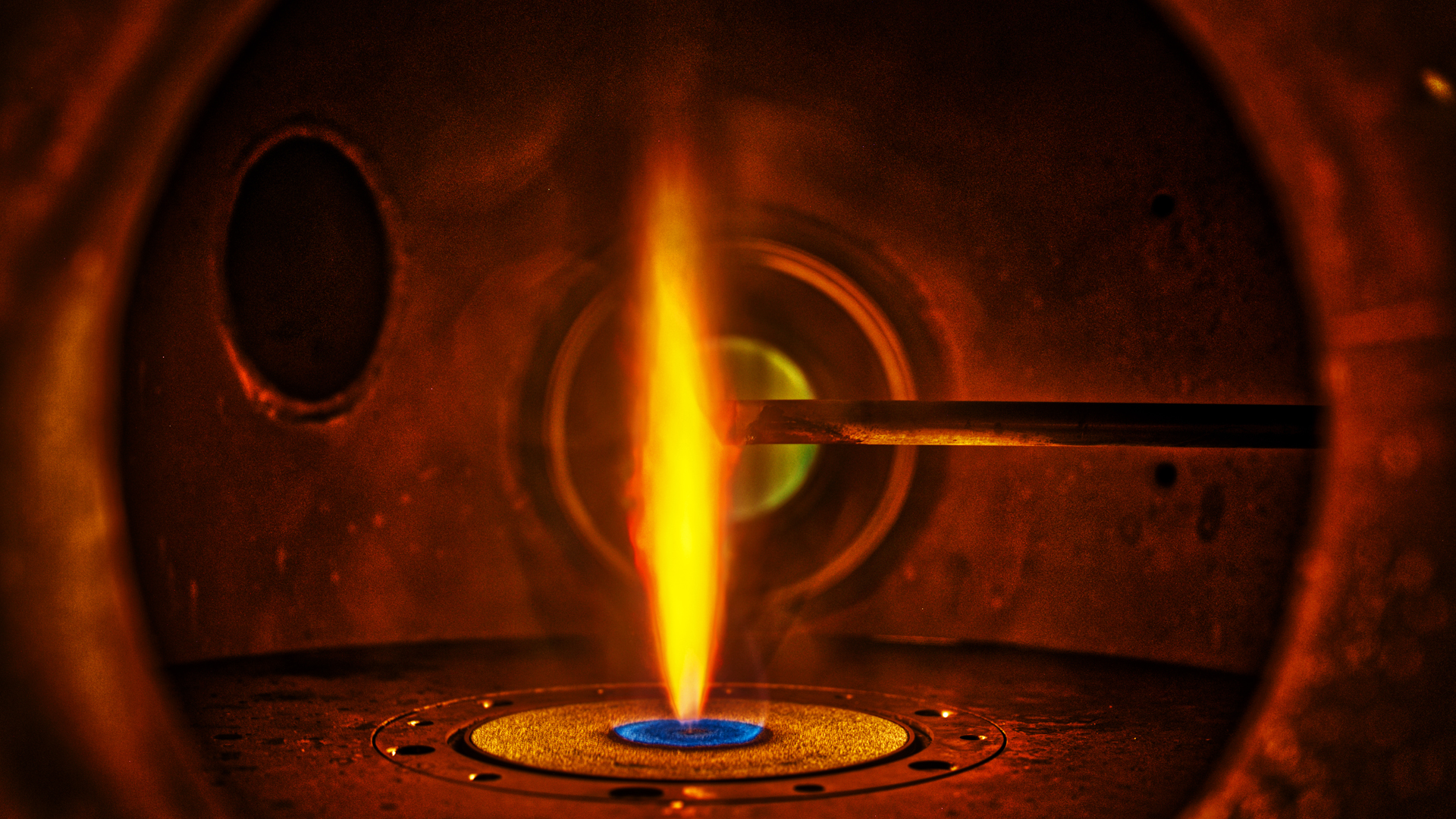 Blick in den Brenner aus Metall: Von unten schießt die Flamme wie aus einem Bunsenbrenner empor. Von rechts schiebt sich ein Sondenstab in die Flamme.