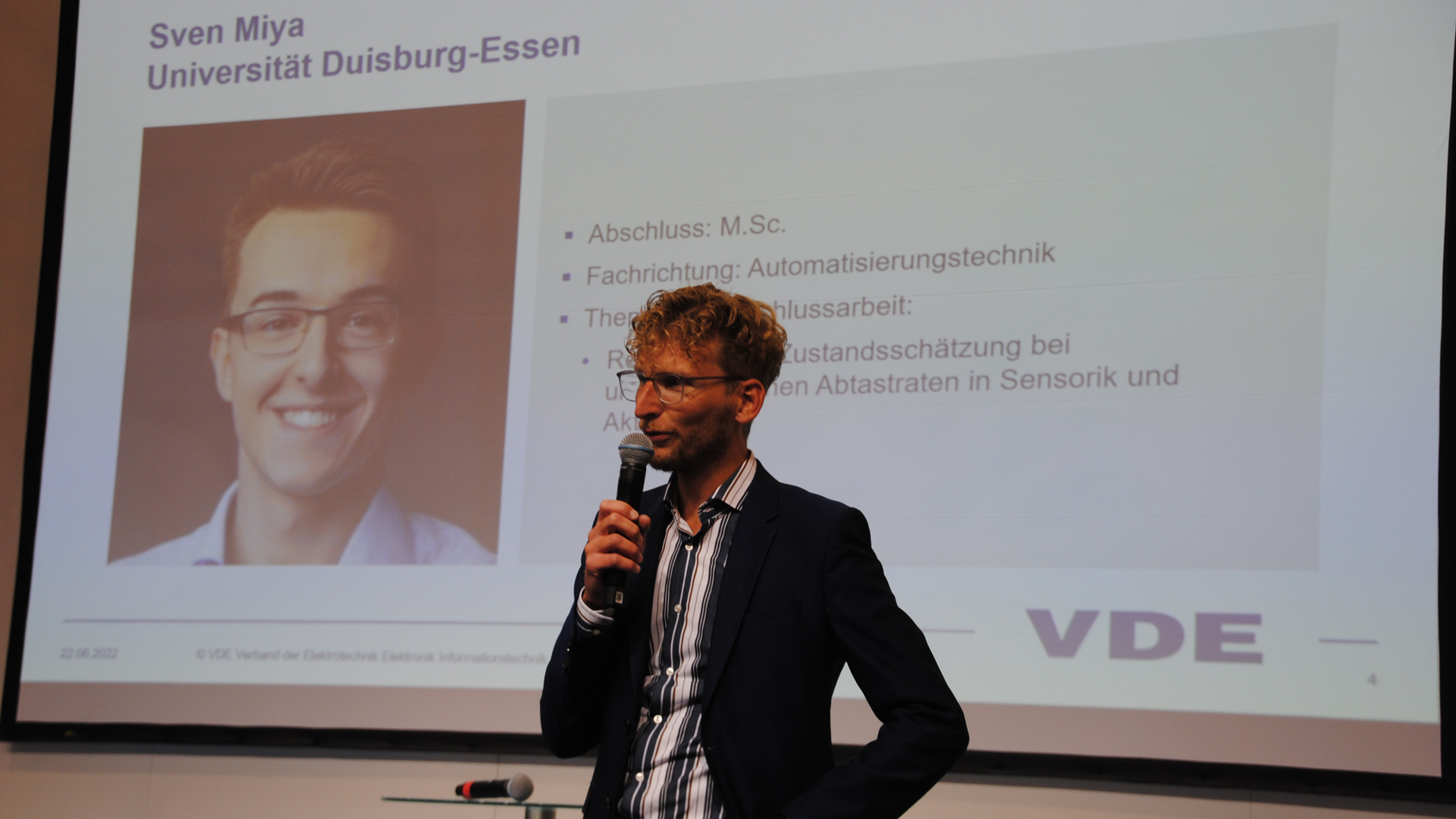 Mann, der in ein Mikrofon spricht. Hinter ihm eine Leinwand, auf der Schrift und ein Protraitbild eine Mannes zu sehen ist.