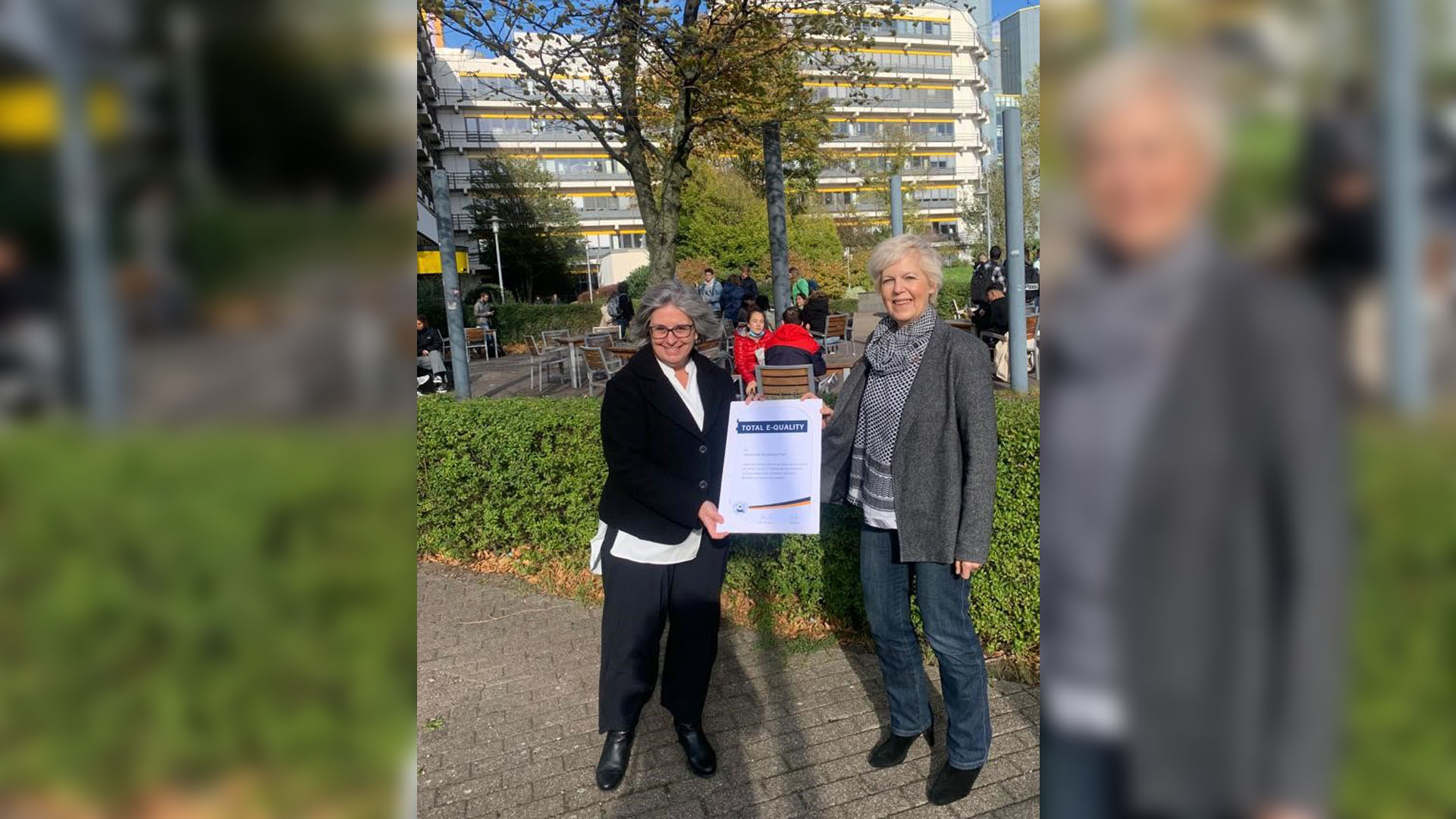 Prof. Shire und Dr. Henze auf dem Essener Campus. Sie halten die Urkunde zwischen sich
