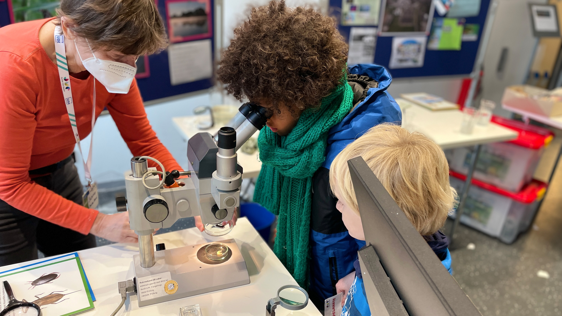 Eine Frau links im Bild zeigt zwei kleinen Kindern wie ein Mikroskop funktioniert