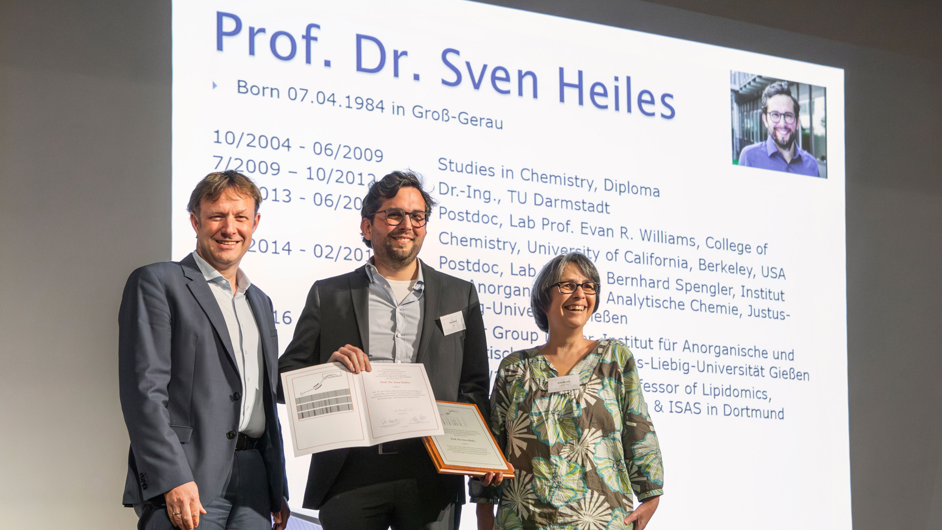 Auf dem Bild sind drei Personen auf einer Bühne zu sehen. In der Mitte steht der Preisträger Sven Heiles und hält seine Urkunde in die Kamera.