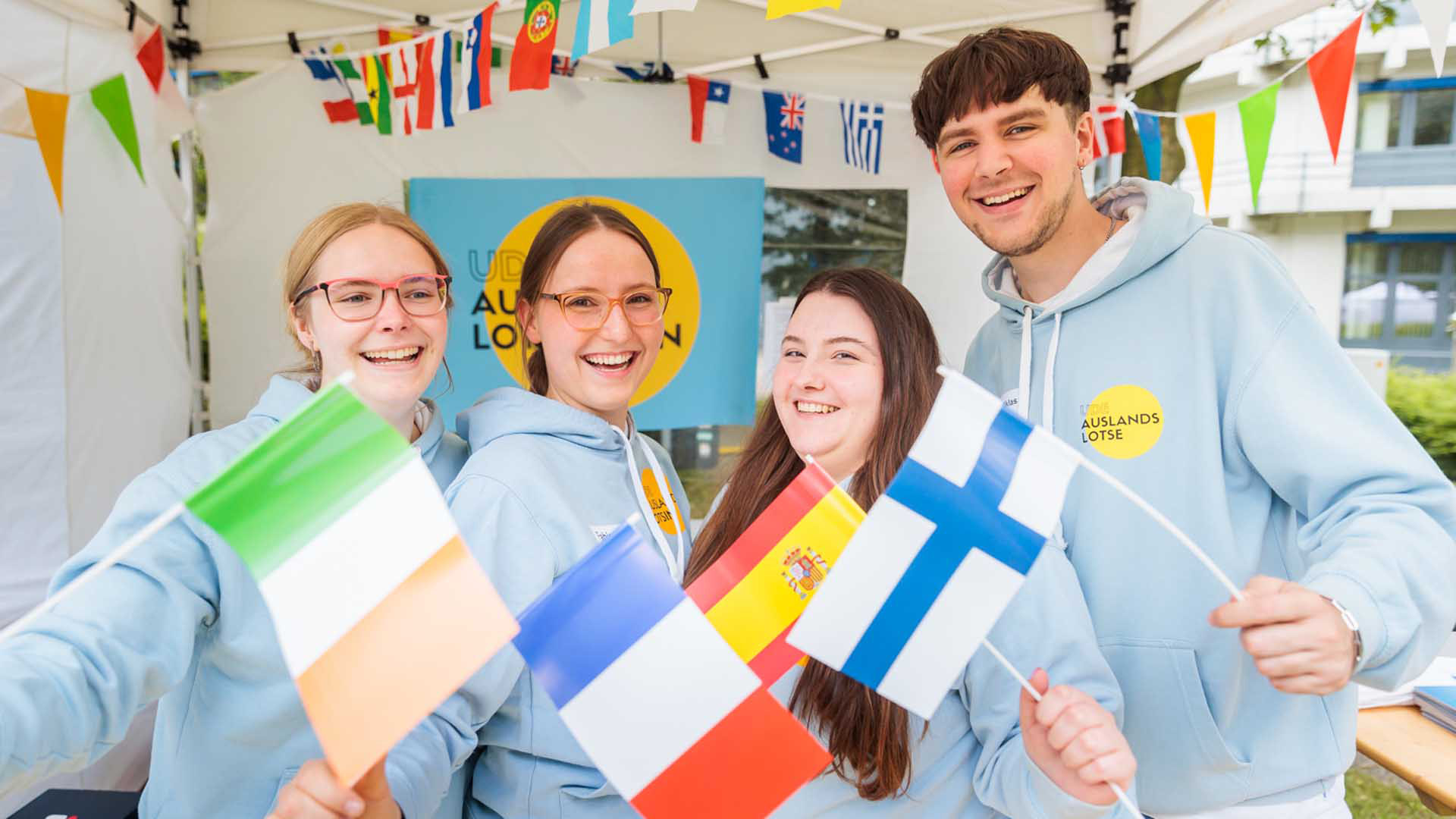 Eine Gruppe von Studierenden (drei Frauen, ein Mann), in einem Pavillon. Sie halten Fähnchen verschiedener Länder in Händen. Die Auslandslotsen präsentierten ihre Arbeit beim letzten UDE-Sommerfest 2022 am Campus Essen.