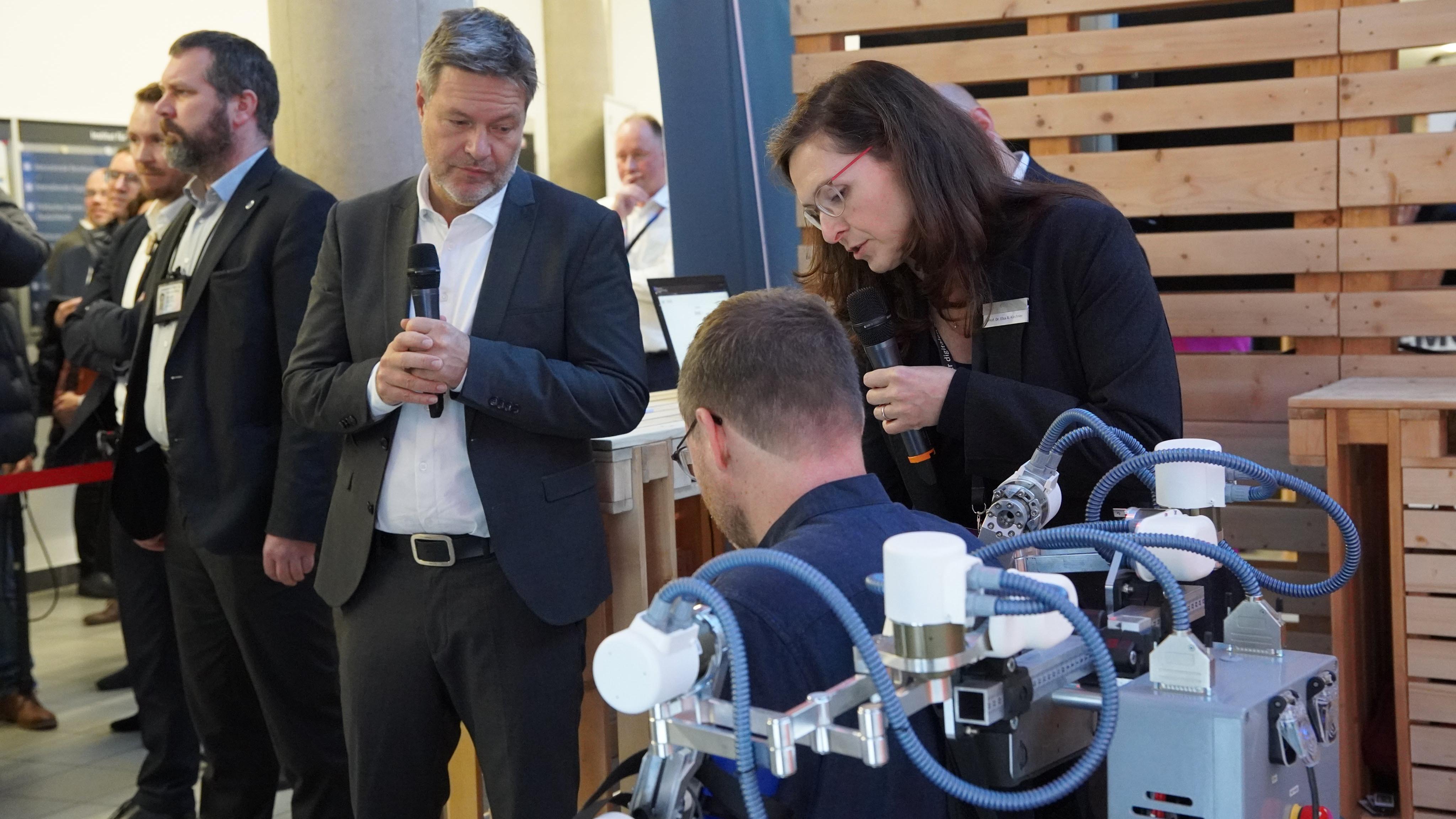 Prof. Elsa Kirchner (UDE) demonstriert Robert Habeck ein robotisches Exoskelett zur Reha von Schlaganfallpatient:innen. Ein Mitarbeiter des DFKI trägt das Exoskelett am Oberkörper zu Demonstrationszwecken