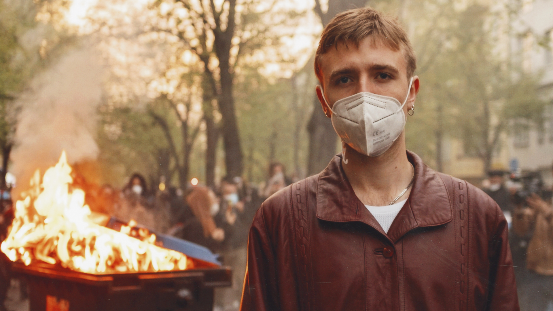 Junger Mann mit Maske vor brennender Mülltonne in Berlin