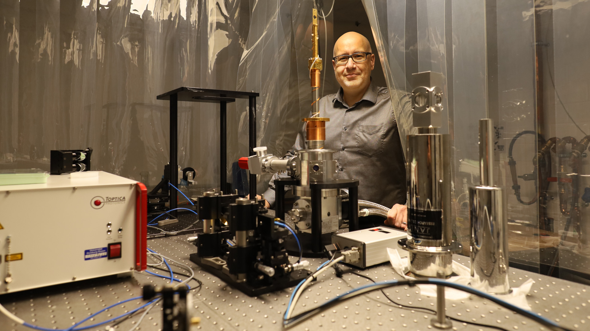 Prof. Mittendorff peeking through an opening in a transparent slatted curtain. He looks over a metal table with a set up of several technical devices.