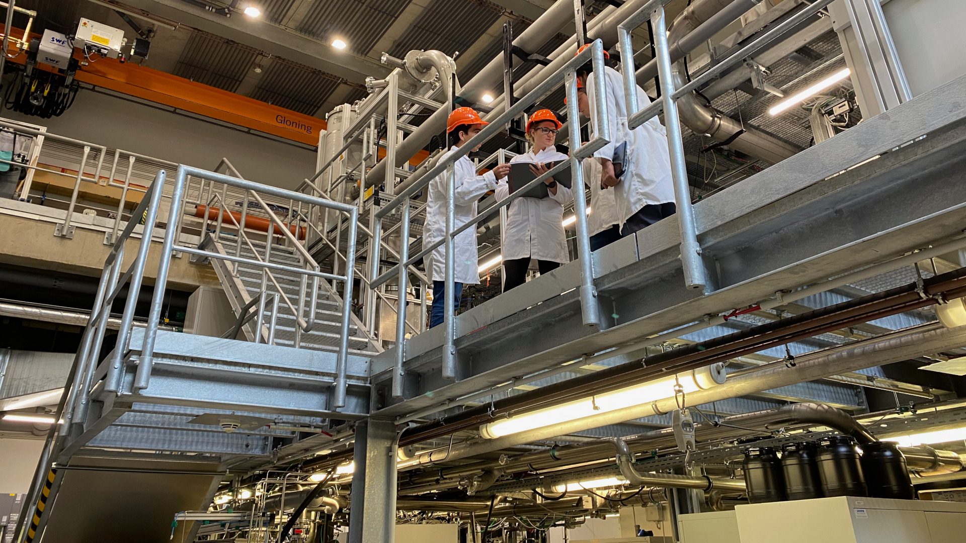 View from the lowest floor to an upper grid level in the multi-storey laboratory of the nanoparticle synthesis facilities. Three scientists in lab coats and helmets are having a discussion.