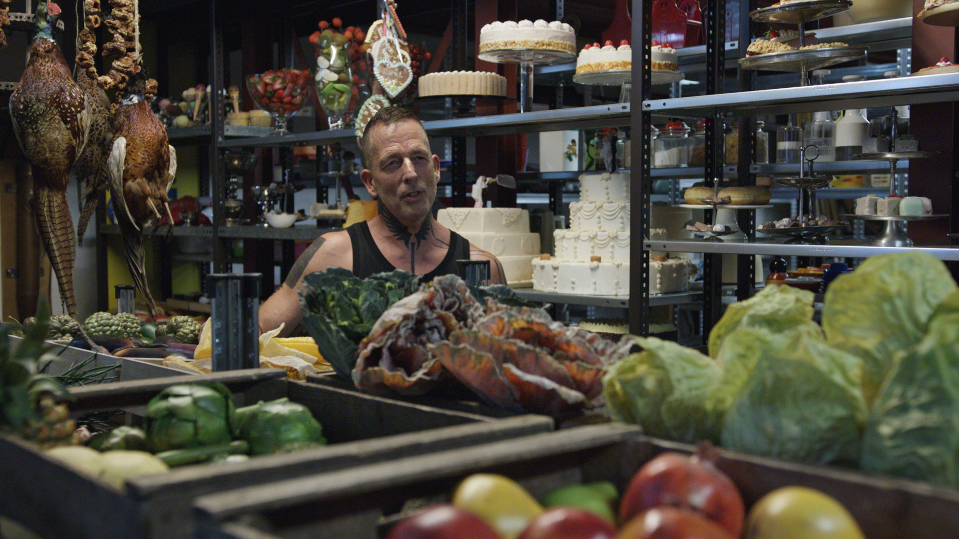 Ein Mann in Muscleshirt und mit vielen Tätowierungen steht inmitten prall gefüllter Regale mit Essens-Requisiten. Vor ihm liegt Gemüse, neben ihm hängen zwei Fasane, im Hintergrund Torten.