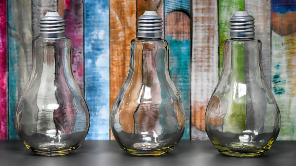 Drei Glühbirnen stehen vor einer bunt angestrichenen Holzwand. Die einzelnen Latten dieser sind unregelmäßig farbig angestrichen, in den Farben blau, rot, orange, grün und Petrol