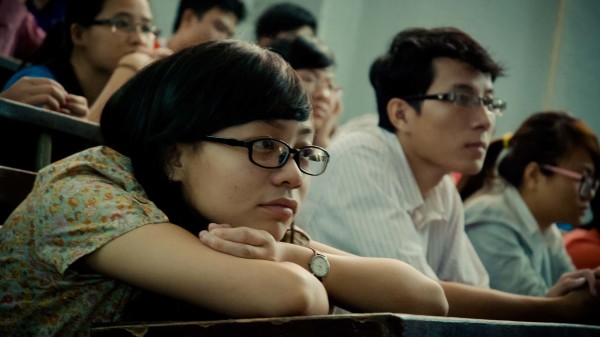 Studierende sitzen in einem Hörsaal. Im Bildausschnitt sitzt eine junge Frau mit auf den Armen aufgestütztem Kopf und blickt nach vorne. Neben ihr und hinter ihr sind weitere Personen zu sehen, die ebenfalls nach vorne schauen.