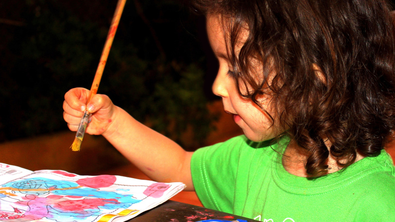 Kind in knallgrünem Shirt mit Pinsel in der Hand, schaut auf ein Bild mit Wasserfarben gemalt