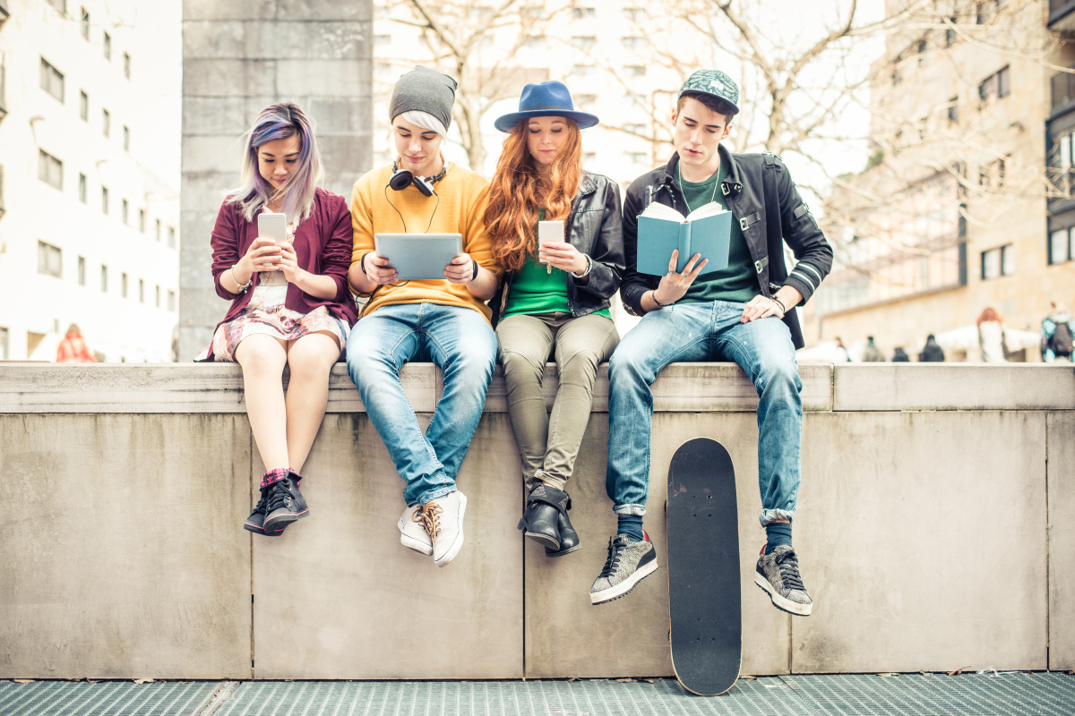 Vier Teenager sitzen auf einer Stufe. Sie haben Handys, Bücher oder Tablets in der Hand.