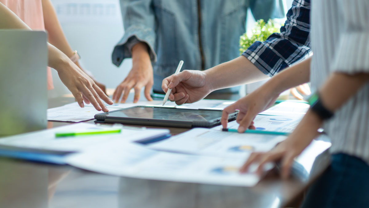 Mehrere Menschen stehen an einem Tisch mit Dokumenten, ihre Gesichter sind nicht im Bild. 