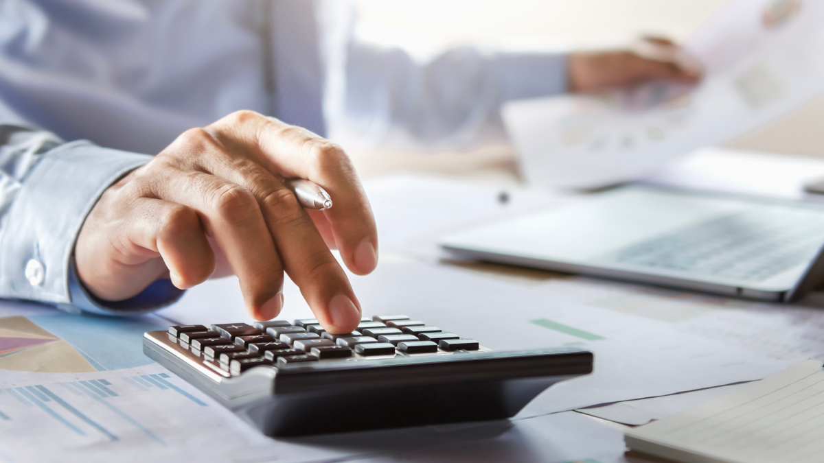 A hand types into a calculator, a laptop and documents on a desk in the background.