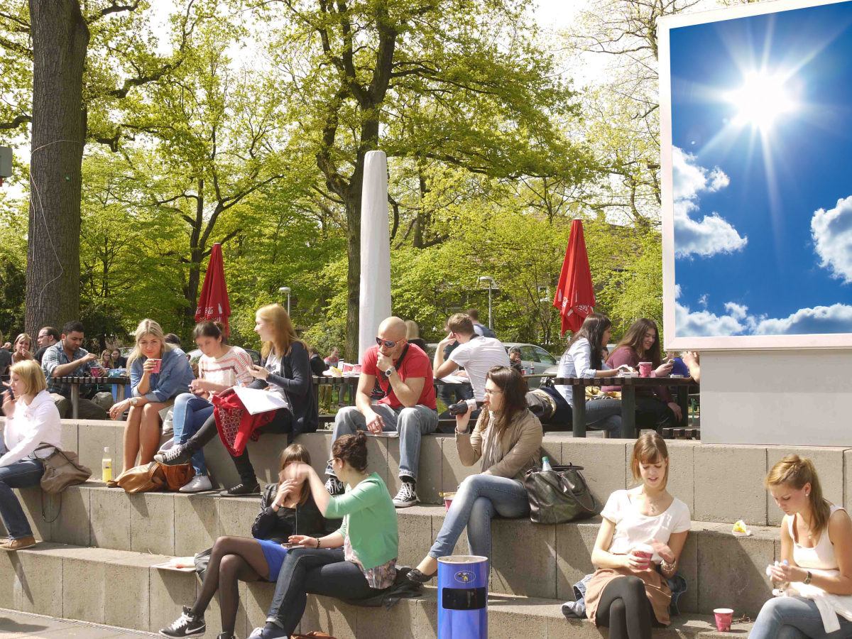 Zu sehen sind Studierende auf dem Campus Essen, die auf Stufen sitzen und die Sonne genießen. Hinter ihnen ist das UDE-Wolkenmotiv zu erkennen.