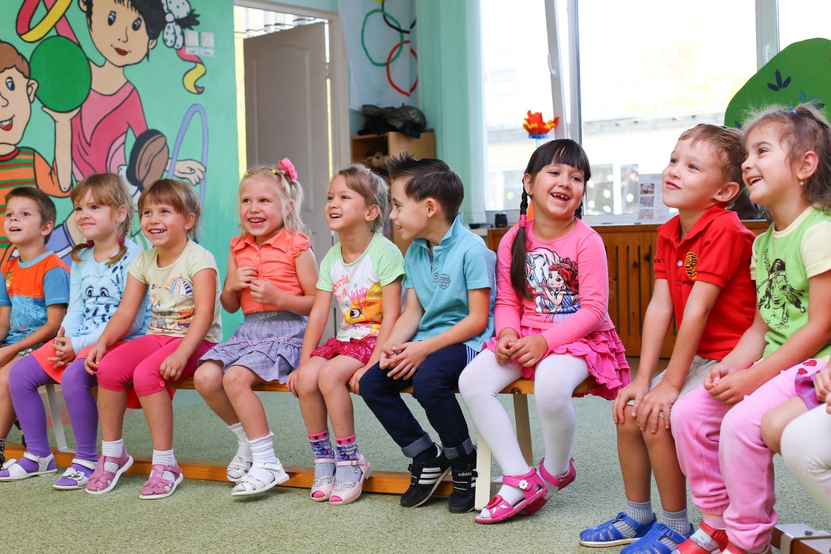(Symbolbild) Es ist eine Gruppe Kinder im Kindergarten zu sehen. Sie sitzen in einer Reihe nebeneinander und lachen.