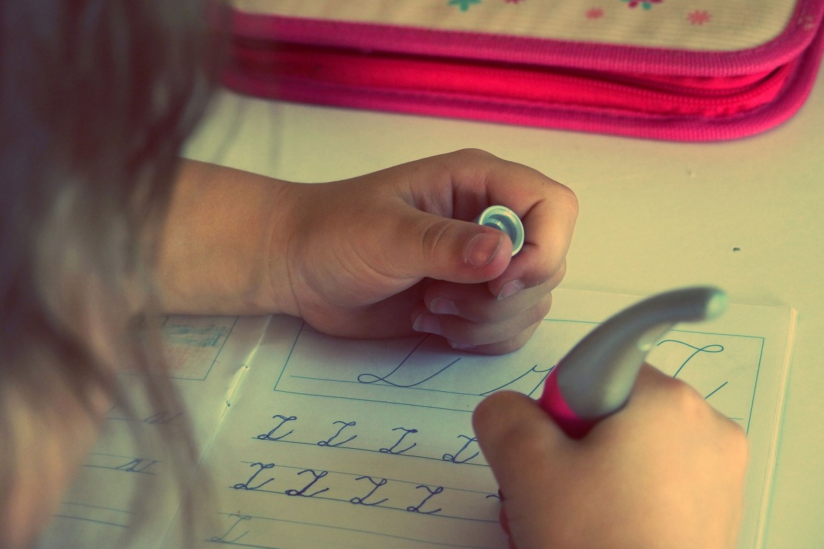 Zu sehen ist ein blondes Mädchen von hinten, das Schreiben übt. Sie hat einen pinken Stift in der Hand.