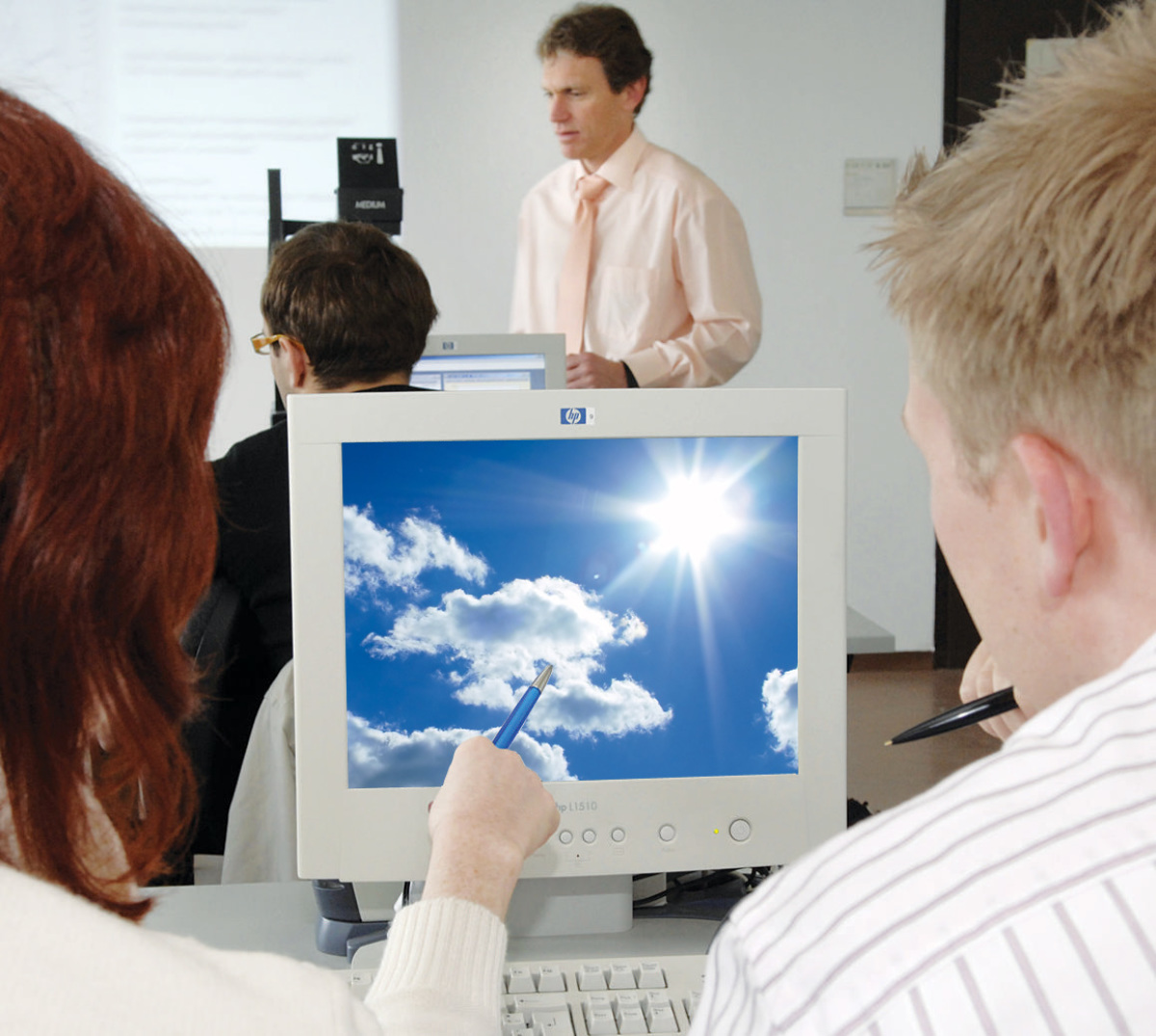 Zu sehen ist eine blonde Frau von hinten, die einem dunkelhaarigen Mann mit einem Stift etwas auf dem Bildschirm vor ihnen zeigt. Der Bildschirm ist mit dem die UDE repräsentierenden Wolkenmotiv ausgefüllt.