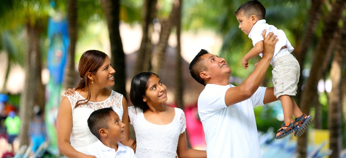 (Symbolbild) eine Familie, bestehend aus Vater, Mutter und drei Kindern, gekleidet in weiß, steht in einer Allee. Der Vater hebt einen Sohn hoch. Alle lächeln oder lachen.