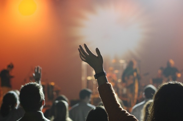 Zu sehen sind Leute auf einem Konzert, die einer Band zujubeln. Im Hintergrund ist ein strahlendes Licht zu sehen. Rechts im Vordergrund hebt eine Frau ihre Arme.