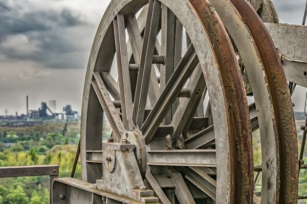 Zu sehen ist ein großes Rad eines alten Stahlwerks. Im Hintergrund lässt sich ein weiteres Stahlwerk und die Bäume des Landschaftsparks Duisburg erkennen.